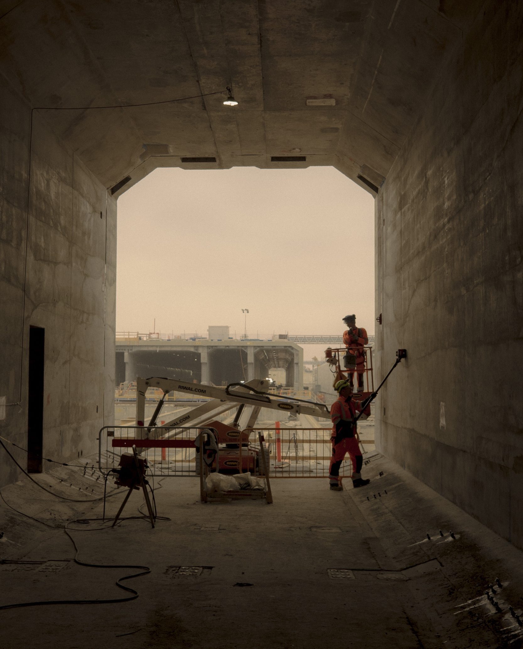 To bygningsarbejdere i sikkerhedsudstyr arbejder på betonvæggen inde i en stor tunnel, med udstyr og barrierer omkring dem og en overskyet byggeplads, der kan ses gennem tunnelindgangen.