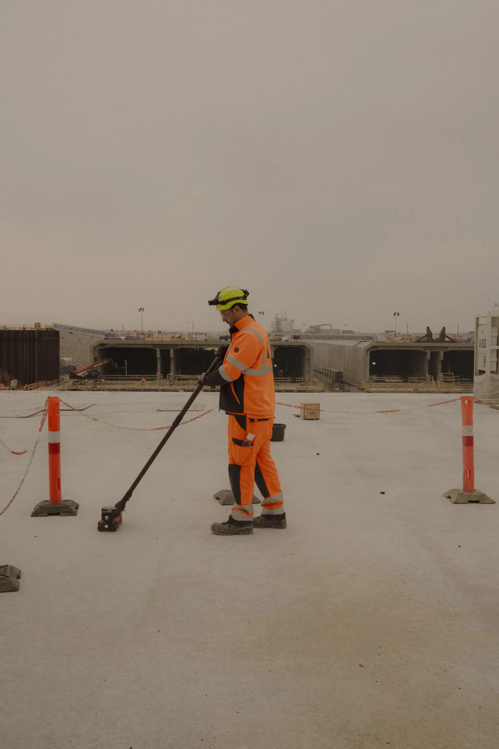 En bygningsarbejder i orange sikkerhedsudstyr og hjelm bruger et værktøj på en betonoverflade på en udendørs byggeplads, hvor man kan se tunneler og orange sikkerhedskegler i baggrunden.