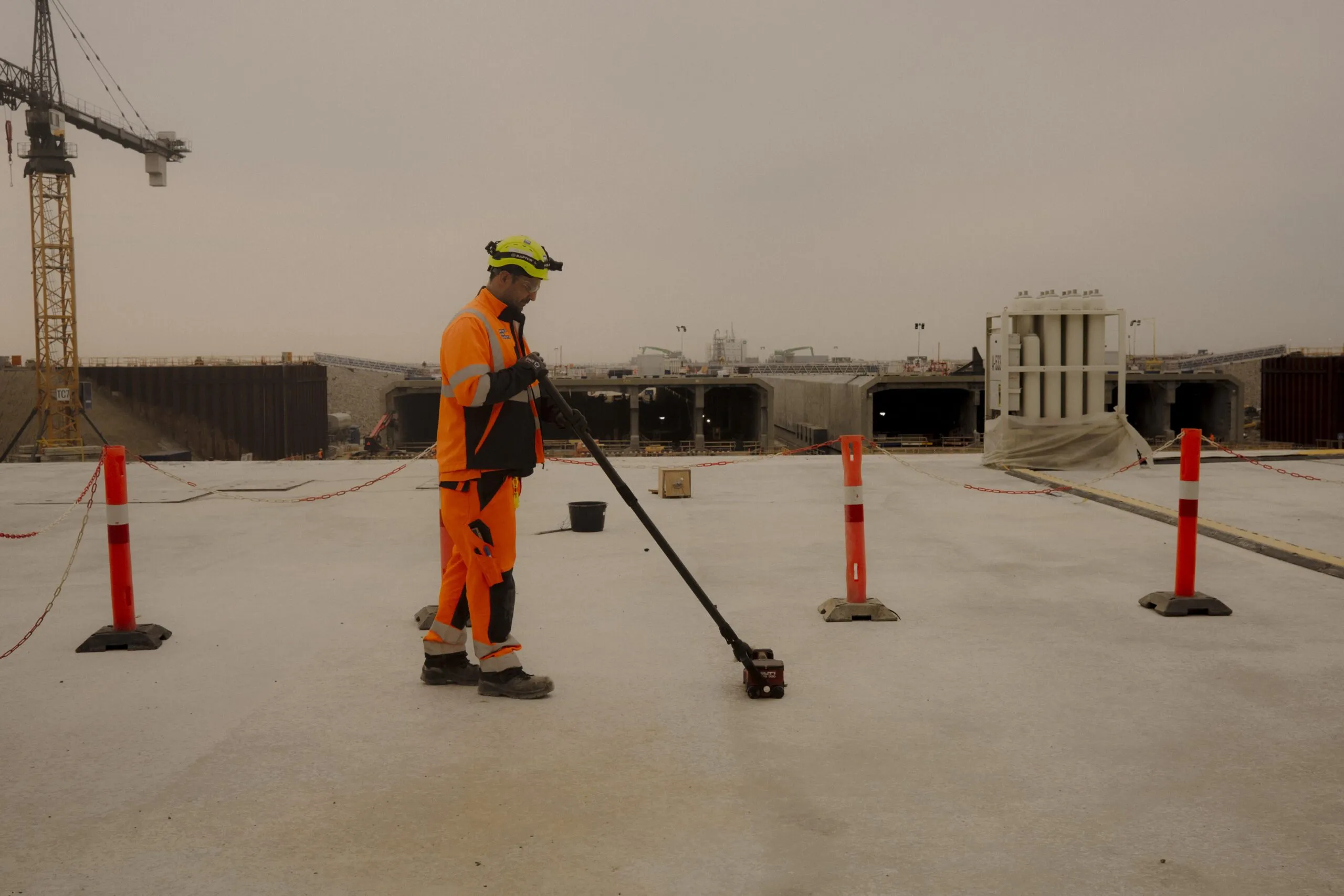 En bygningsarbejder i orange sikkerhedsudstyr bruger et målehjul på en betonoverflade på en byggeplads med kraner og ufærdige bygninger i baggrunden. Orange sikkerhedskegler markerer arbejdsområdet.