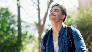 En ung mand med krøllet hår og rygsæk kigger op og smiler, mens han står udendørs omgivet af træer og sollys.