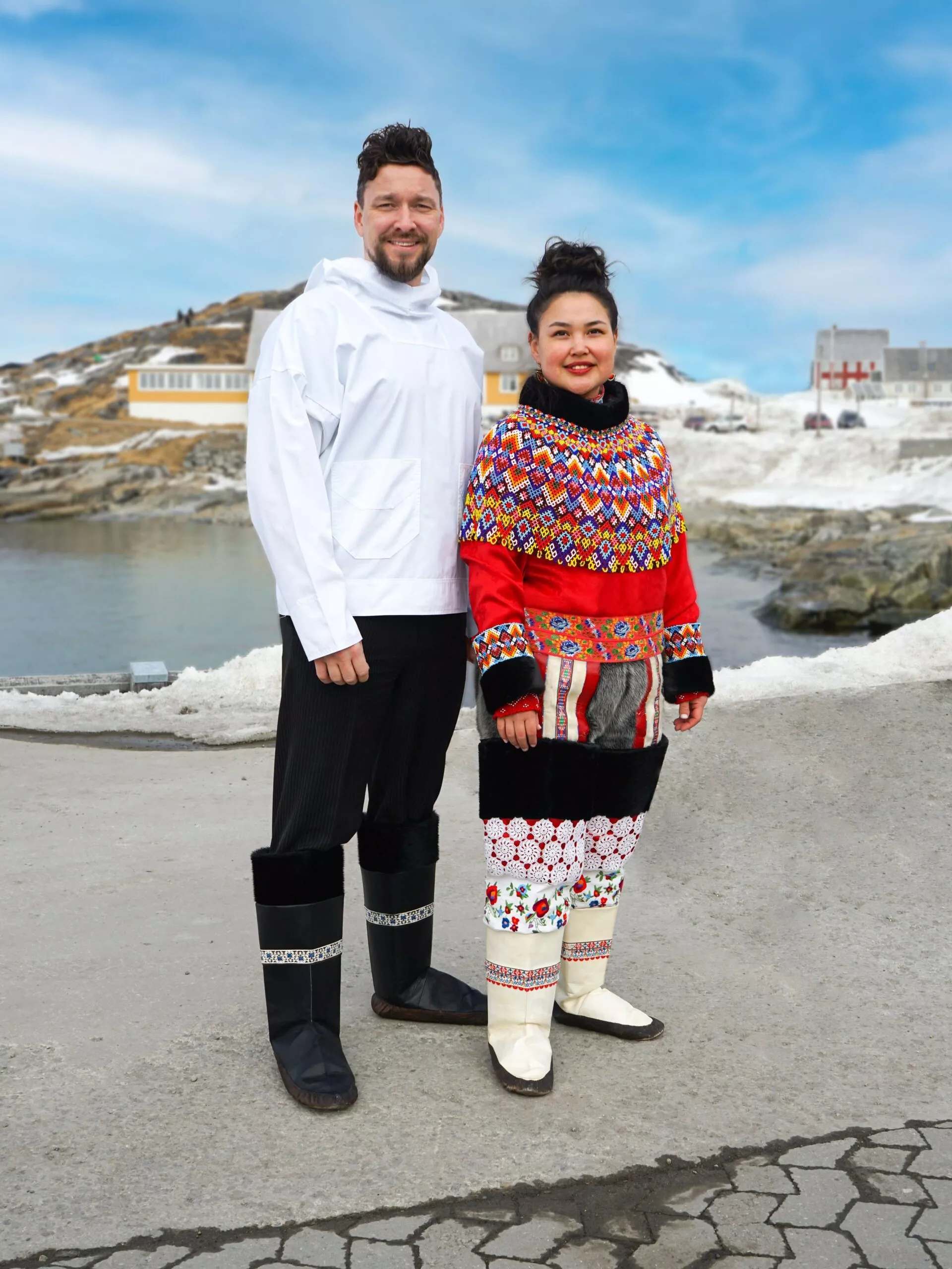 En mand og en kvinde står udendørs på en snedækket gade, begge iført traditionelt grønlandsk tøj med farverige mønstre og pelsdetaljer. I baggrunden ses huse og iskoldt vand under en blå himmel.
