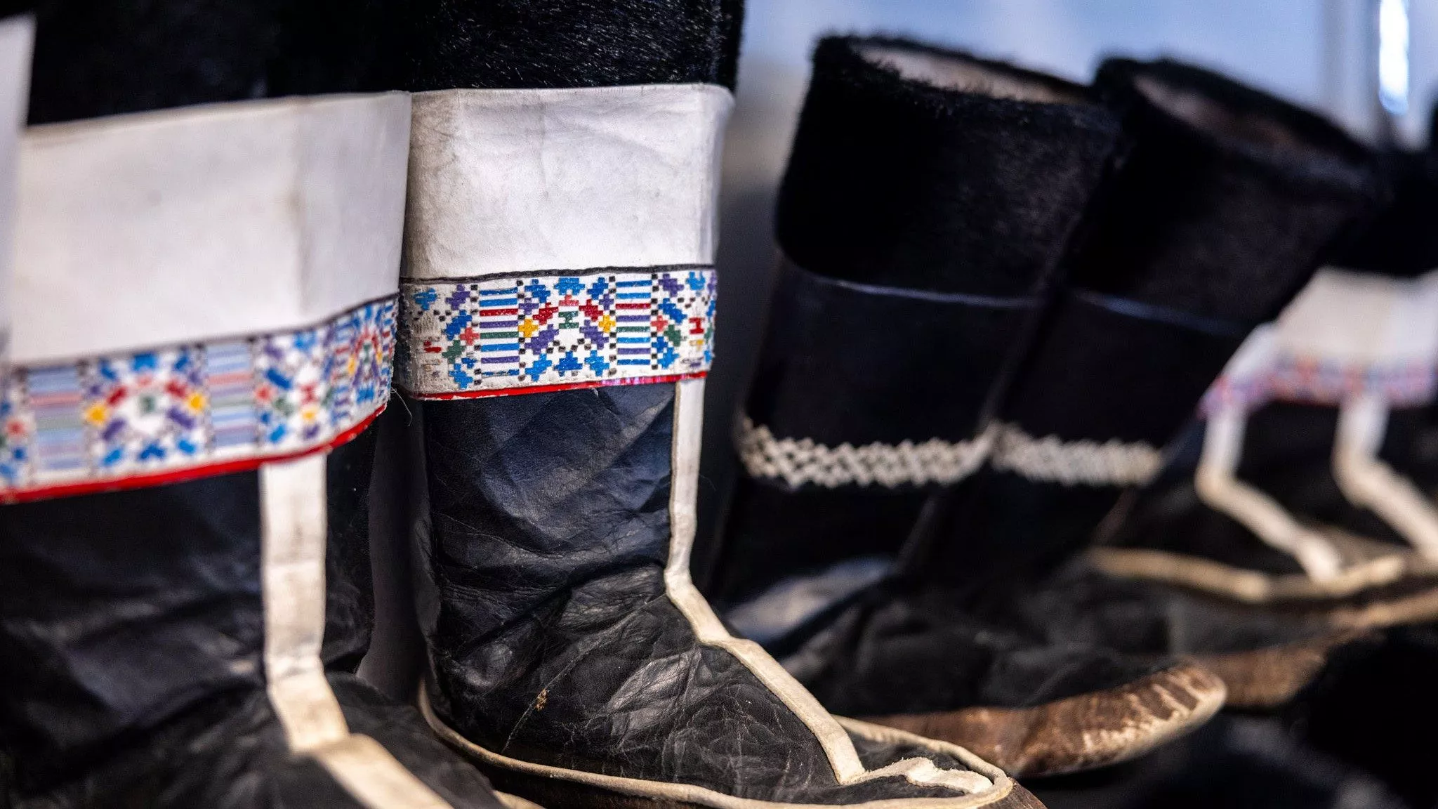 En række traditionelle sorte støvler med dekorativt perlearbejde og mønstre på manchetterne, med indviklede, farverige mønstre på nogle par. Støvlerne ser ud til at være håndlavede og er stillet tæt op ad hinanden.