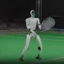 En humanoid robot står på en indendørs tennisbane med en tennisketcher i hånden og ser ud til at være klar til at spille. Omgivelserne består af kunstgræs og bænke langs siden.
