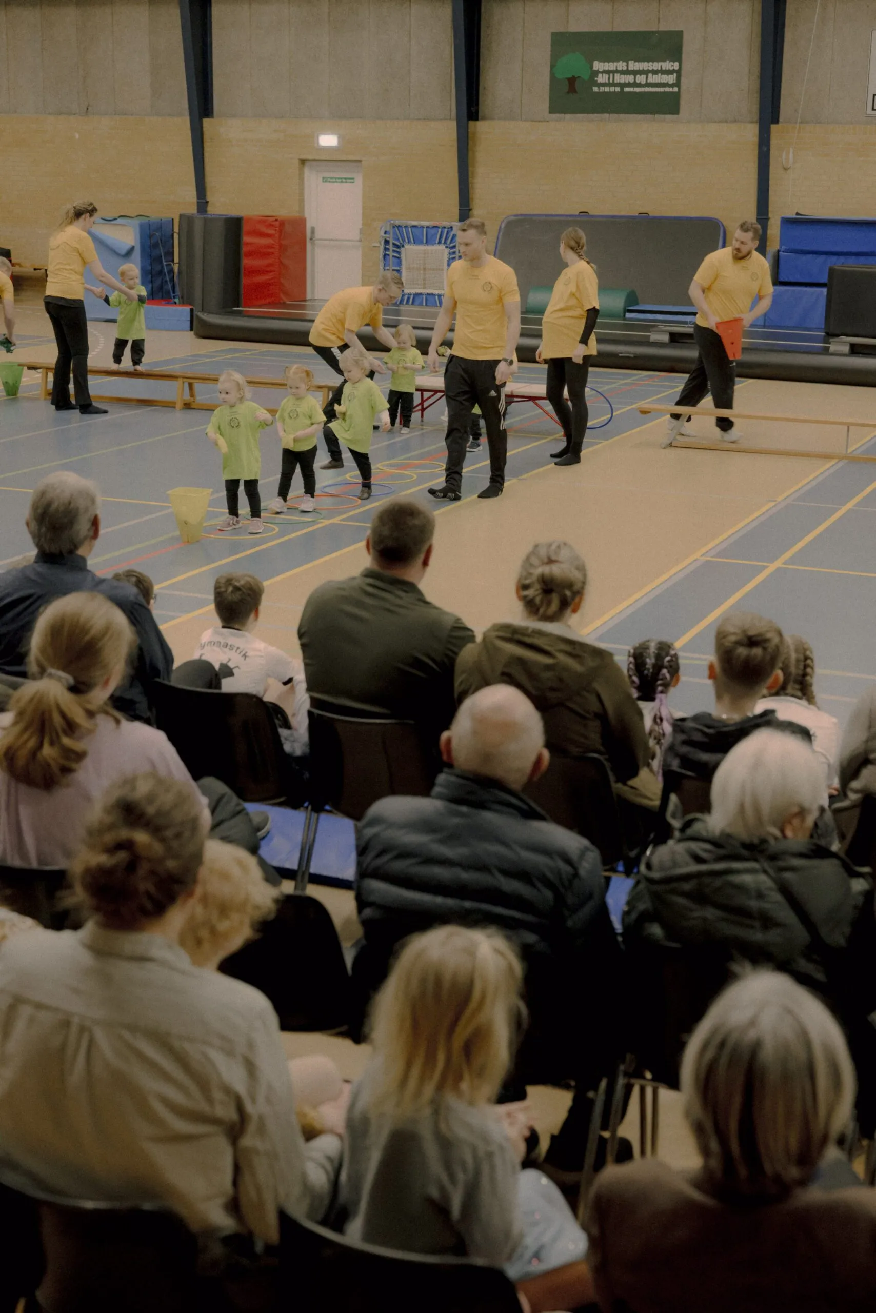 En gruppe små børn og voksne, alle i sportstøj, deltager i en aktivitet i en gymnastiksal, mens et publikum af siddende voksne og børn ser på fra forgrunden.