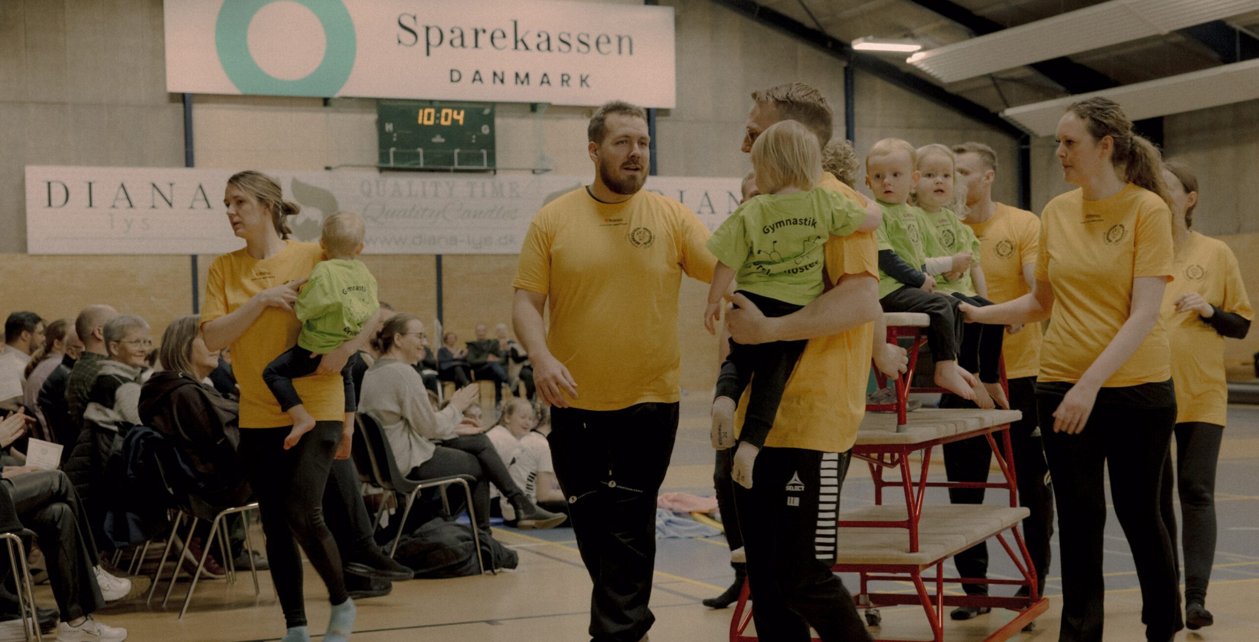 Voksne og små børn i gule og grønne skjorter går og bærer børn på et gymnastiksalsgulv, med tilskuere siddende i baggrunden og et Sparekassen Danmark-skilt over hovedet.