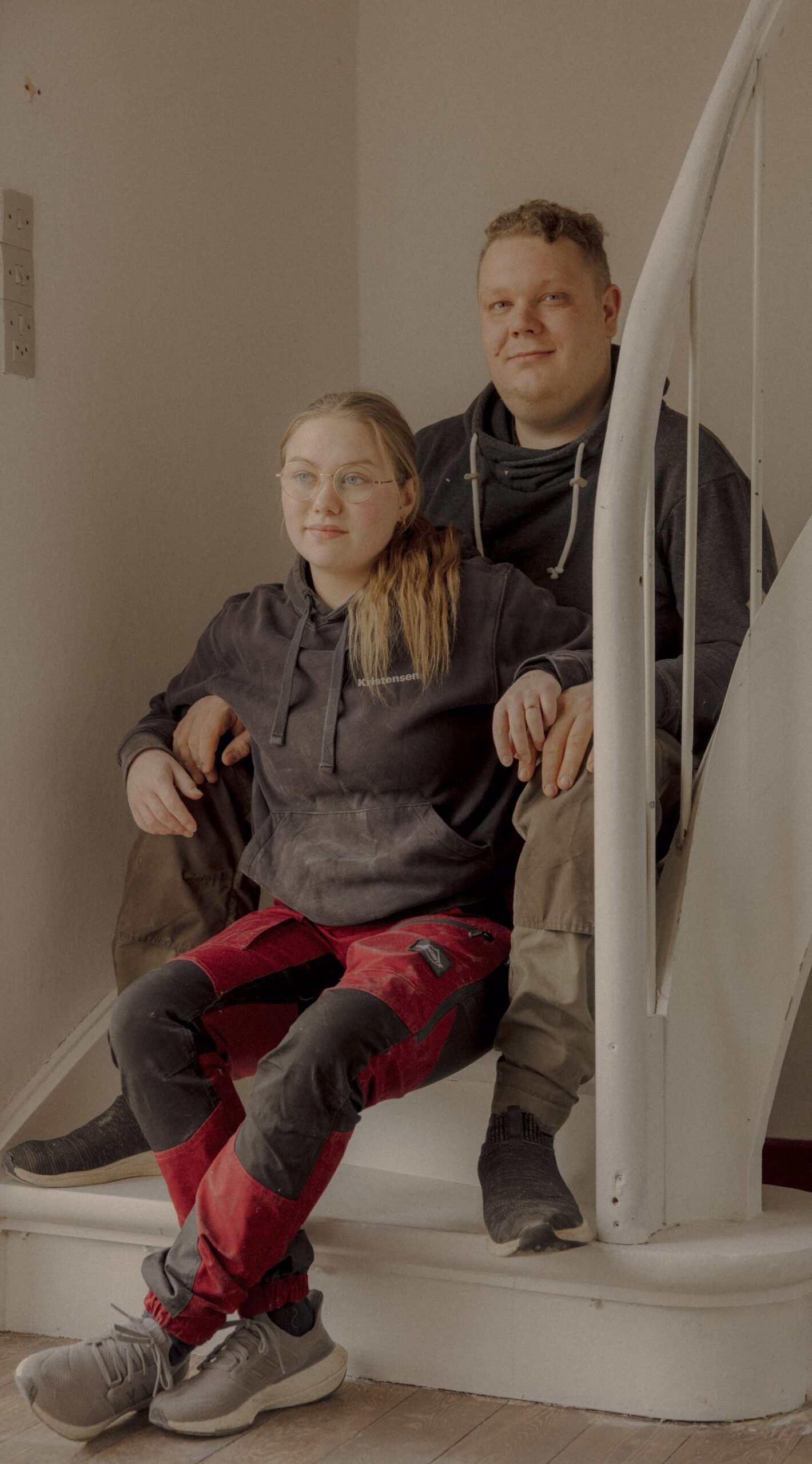 To personer i arbejdstøj sidder sammen på en hvid vindeltrappe i et svagt oplyst rum. Personen foran har blondt hår og briller; personen bagved smiler med en arm omkring sig. Omgivelserne virker afslappede og uformelle.