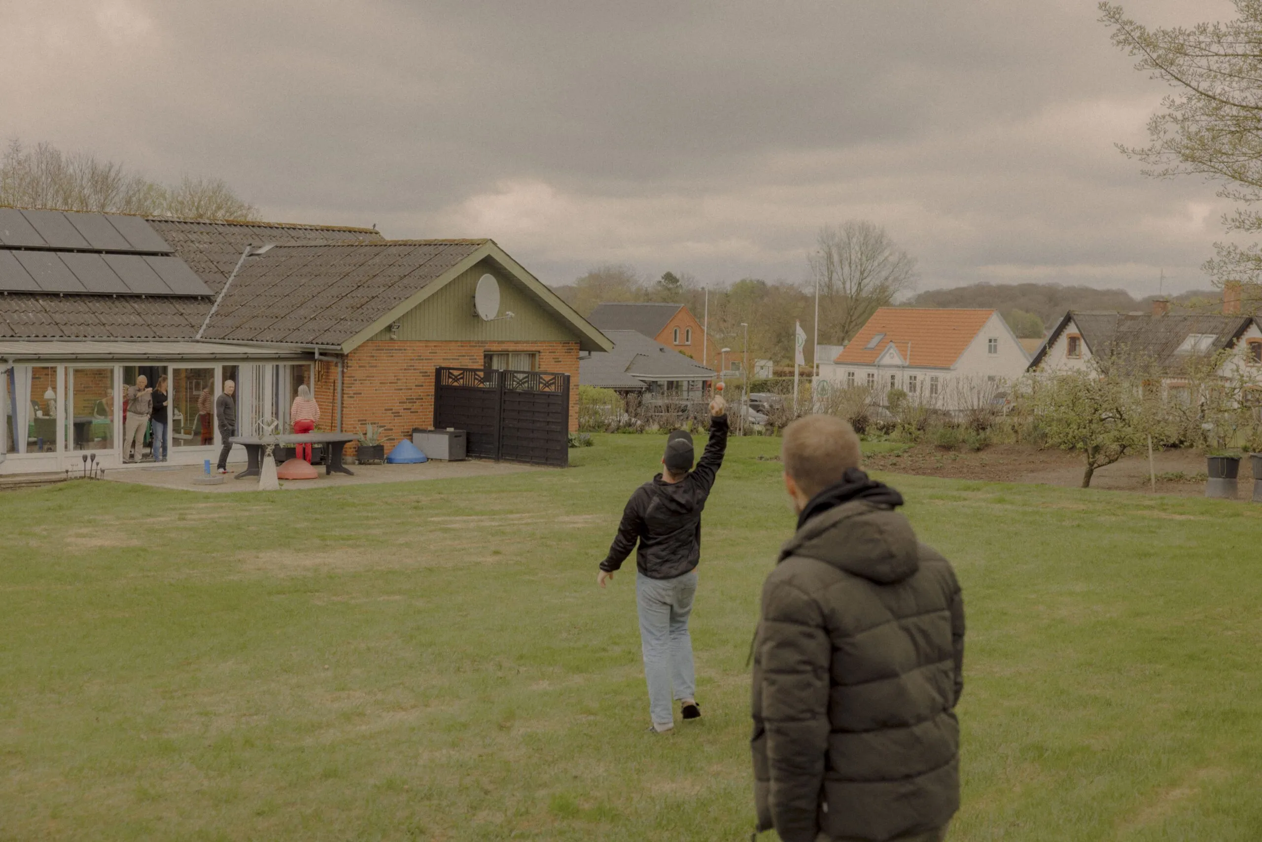 To personer står uden for et hus på en græsplæne; den ene løfter en arm, mens andre samles i nærheden af huset, som har solpaneler på taget. Himlen er overskyet, og flere huse er synlige i baggrunden.