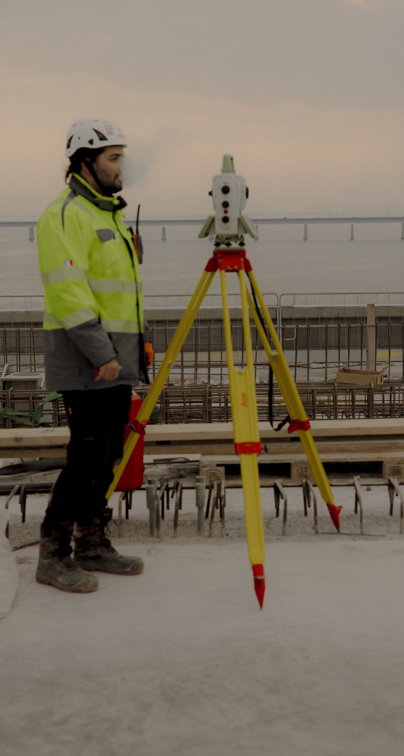 En bygningsarbejder i gul refleksjakke og hjelm står ved siden af et gult opmålingsinstrument monteret på et stativ på en betonplads nær vand, og i baggrunden ses en bro.