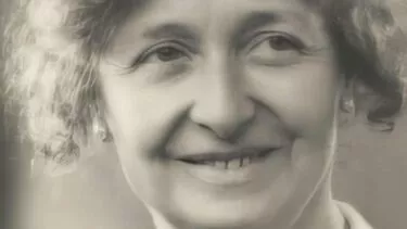 Et sort-hvidt portræt af en ældre kvinde med krøllet hår, som smiler blidt. Hun er iført en lys bluse og en halskæde, og baggrunden er let sløret.