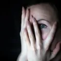 En person dækker det meste af ansigtet med sine hænder og kigger gennem fingrene med et stort, frygtsomt øje. Baggrunden er mørk, hvilket fremhæver udtrykket af angst eller frygt.