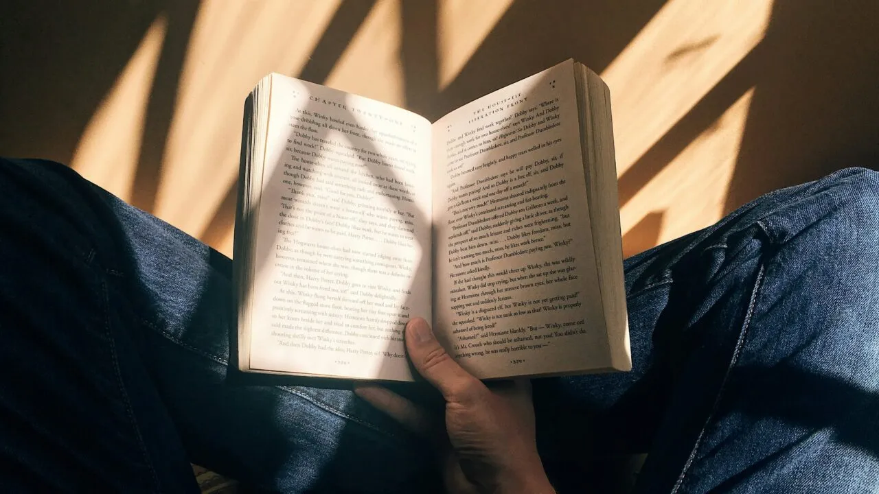 En person iført blå jeans sidder med benene over kors og en åben bog i skødet. Sollys og skygger falder over siderne og personens hånd og skaber en varm og fredfyldt atmosfære.