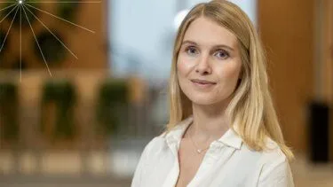 En ung kvinde med langt blondt hår og en hvid skjorte smiler blidt til kameraet. Baggrunden er sløret og viser indendørs planter og varm belysning.