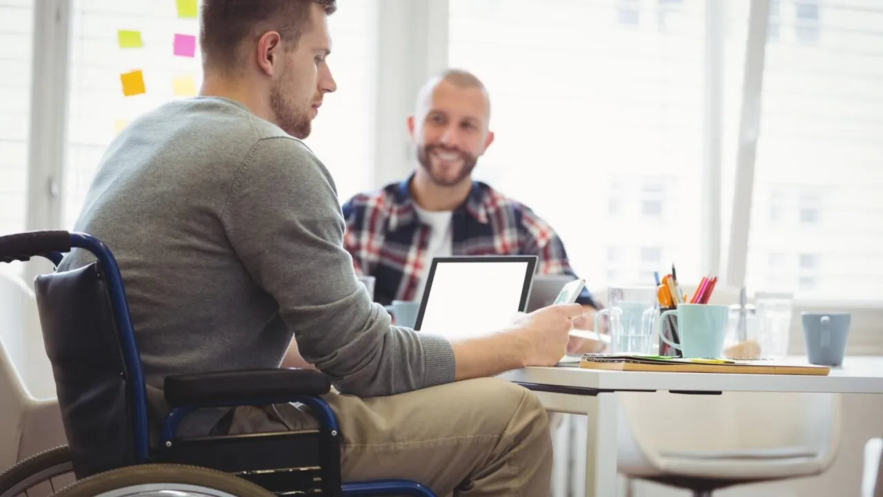 To mænd sidder ved et skrivebord på et kontor, den ene i kørestol, og fører en samtale. De er omgivet af papirer, en tablet og kontorartikler, med lyse vinduer og klistermærker i baggrunden.