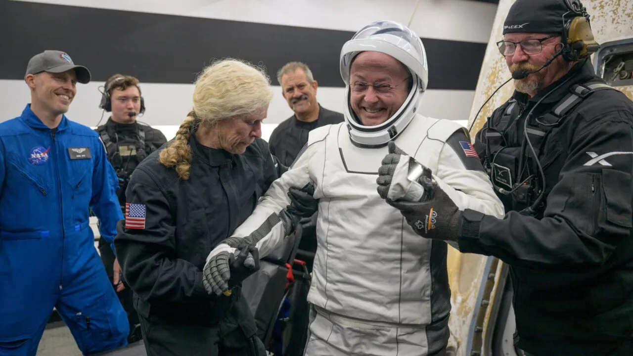 En smilende astronaut i en hvid rumdragt får hjælp af SpaceX- og NASA-besætningsmedlemmer efter at have forladt et rumfartøj, omgivet af folk i uniformer, inde på et anlæg.