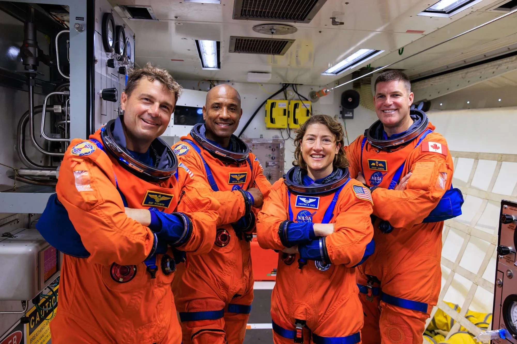 Fire astronauter iført orange NASA-rumdragter står side om side inde i et rumfartøj og smiler med armene over kors. Baggrunden viser teknisk udstyr og kontrolpaneler.