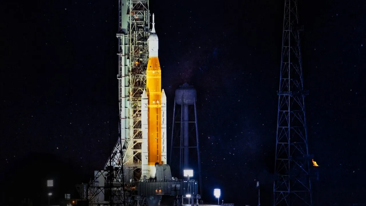 En stor orange og hvid NASA-raket er oplyst på sin affyringsrampe om natten, omgivet af støttestrukturer og tårne, med en mørk, stjernefyldt himmel i baggrunden.