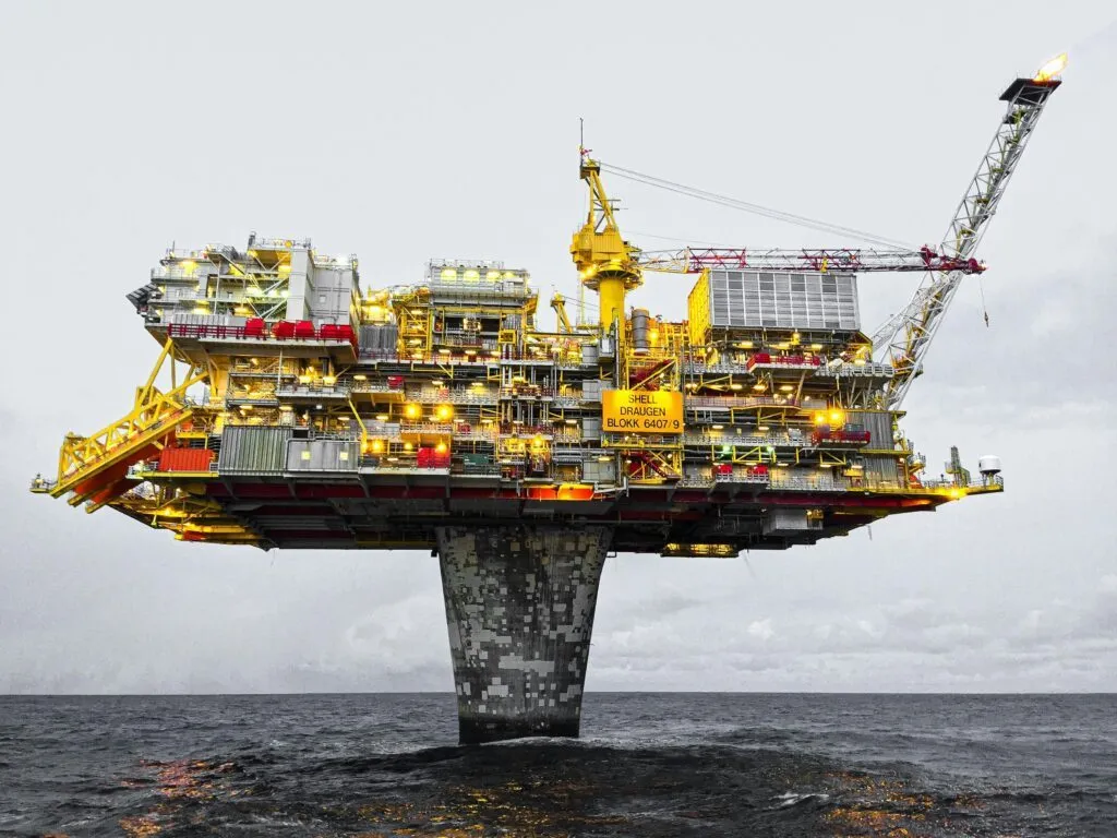 En offshore olieplatform står over mørkt havvand under en overskyet himmel. Strukturen er stærkt oplyst med gule og røde komponenter og en stor kran, der strækker sig udad.