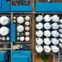Luftfoto af et industrianlæg med mange store, cylindriske lagertanke og bygninger med blåt tag arrangeret i et gitterlignende mønster. Stier og køretøjer er synlige omkring strukturerne.