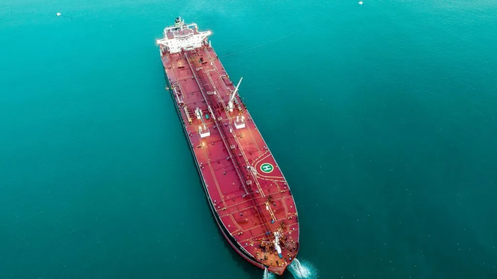 Luftfoto af et stort rødt fragtskib med helikopterlandingsplads, der ligger for anker i roligt, turkisblåt vand.