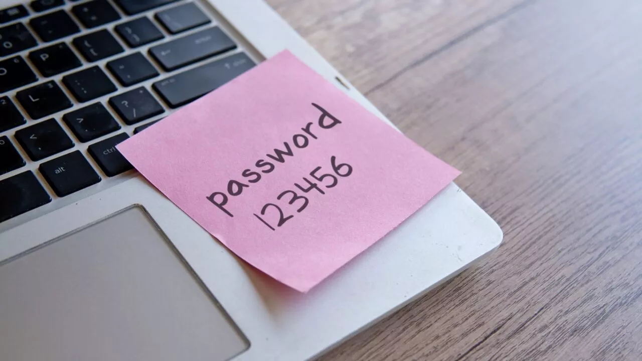 En lyserød seddel med adgangskoden 123456 er fastgjort til et tastatur på en bærbar computer, der står på et træbord.