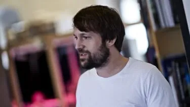 En mand med mellemlangt brunt hår og skæg, iført en hvid T-shirt, står indendørs i nærheden af hylder med bøger og taler eller gestikulerer med sin højre hånd. Baggrunden er let sløret.