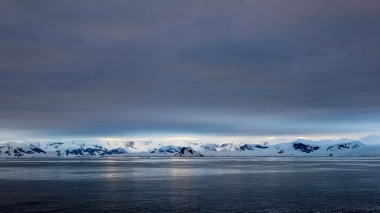 Snedækkede bjerge ligger i horisonten under en dramatisk, overskyet himmel, og deres refleksioner er svagt synlige på det rolige, mørke vand i forgrunden.