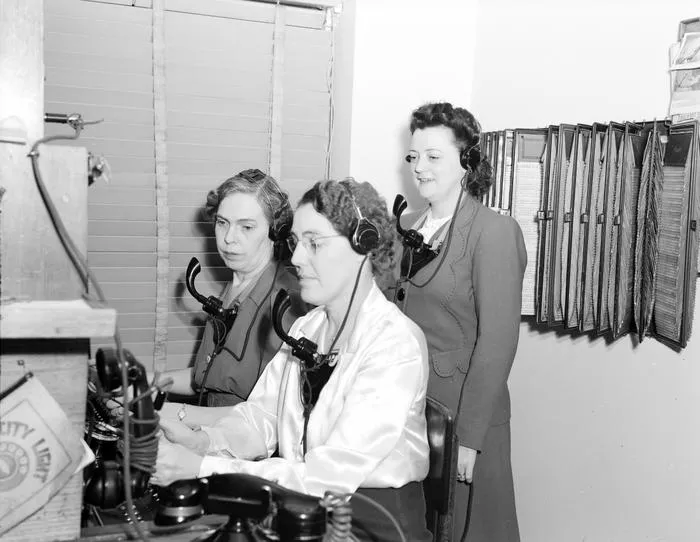 Tre kvinder i vintagetøj betjener et telefonanlæg, to sidder med headset og arbejder, mens en står bag dem og observerer. Omgivelserne ser ud til at være et kontor med udstyr og en mappeorganisation på væggen.