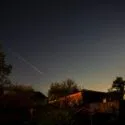 Nattehimmel med mange stjerner, et synligt stjerneskud eller en meteor, der strækker sig diagonalt, og silhuetterne af huse, træer og et hegn i forgrunden.
