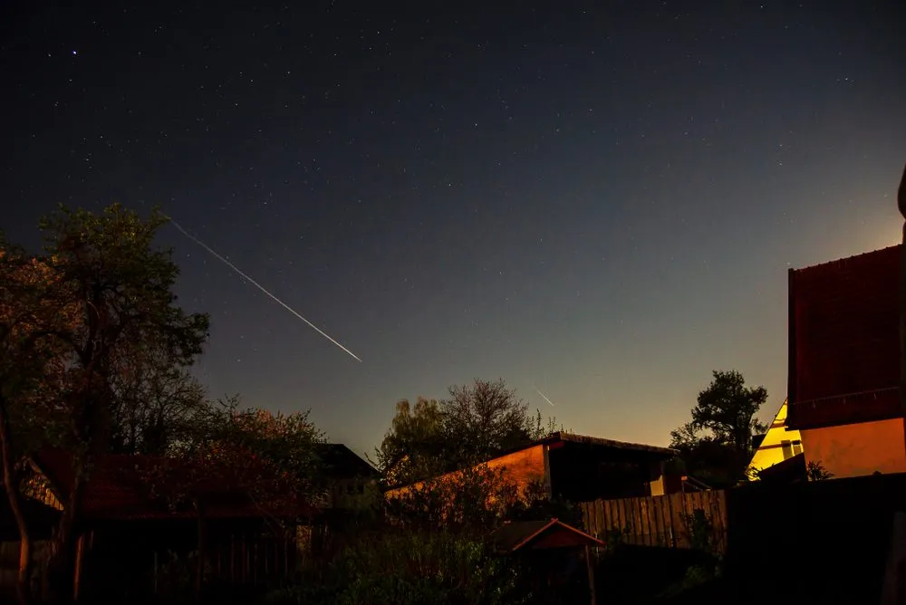 Nattehimmel med mange stjerner, et synligt stjerneskud eller en meteor, der strækker sig diagonalt, og silhuetterne af huse, træer og et hegn i forgrunden.