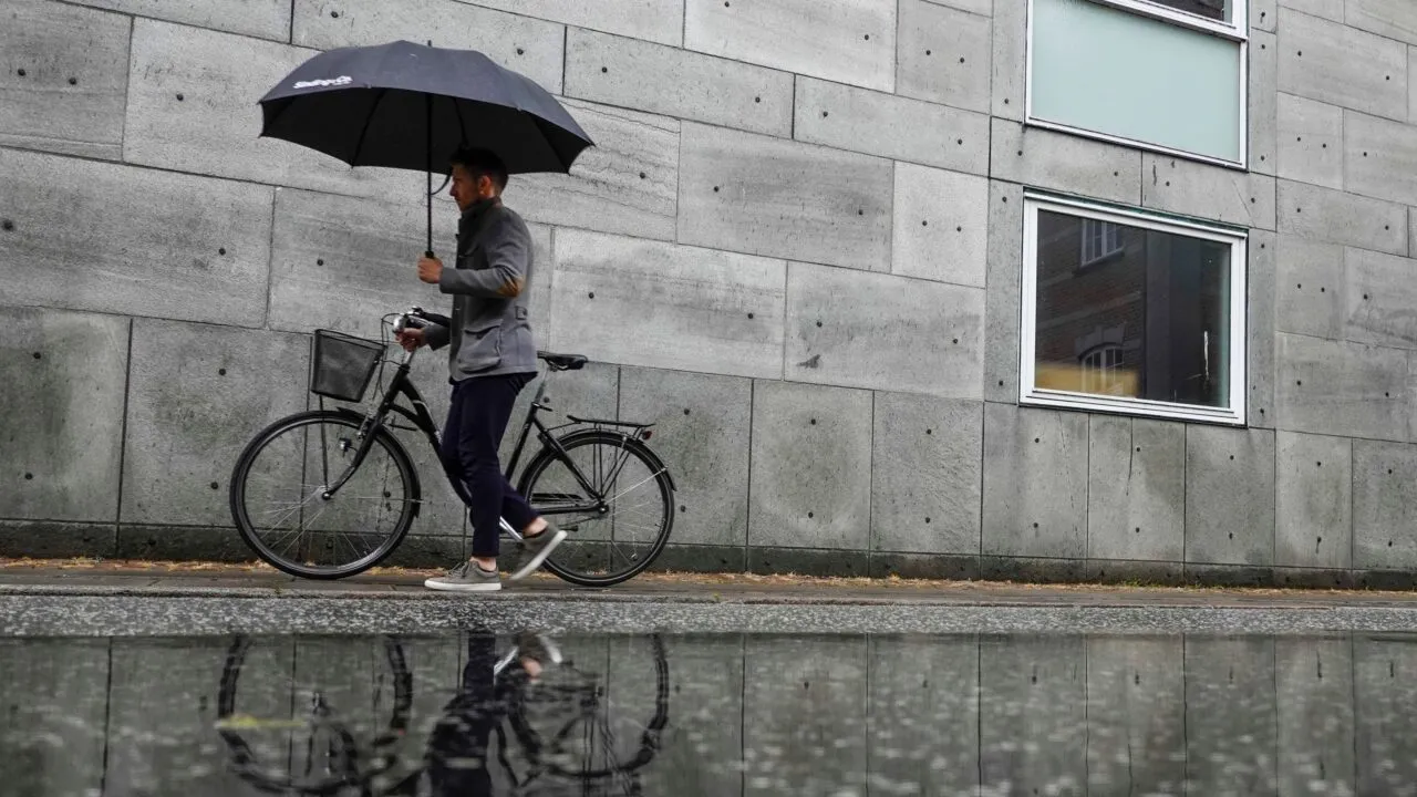En mand går ved siden af en cykel, mens han holder en sort paraply på en regnvejrsdag, og hans spejlbillede er synligt i en stor vandpyt ved en moderne grå betonbygning.