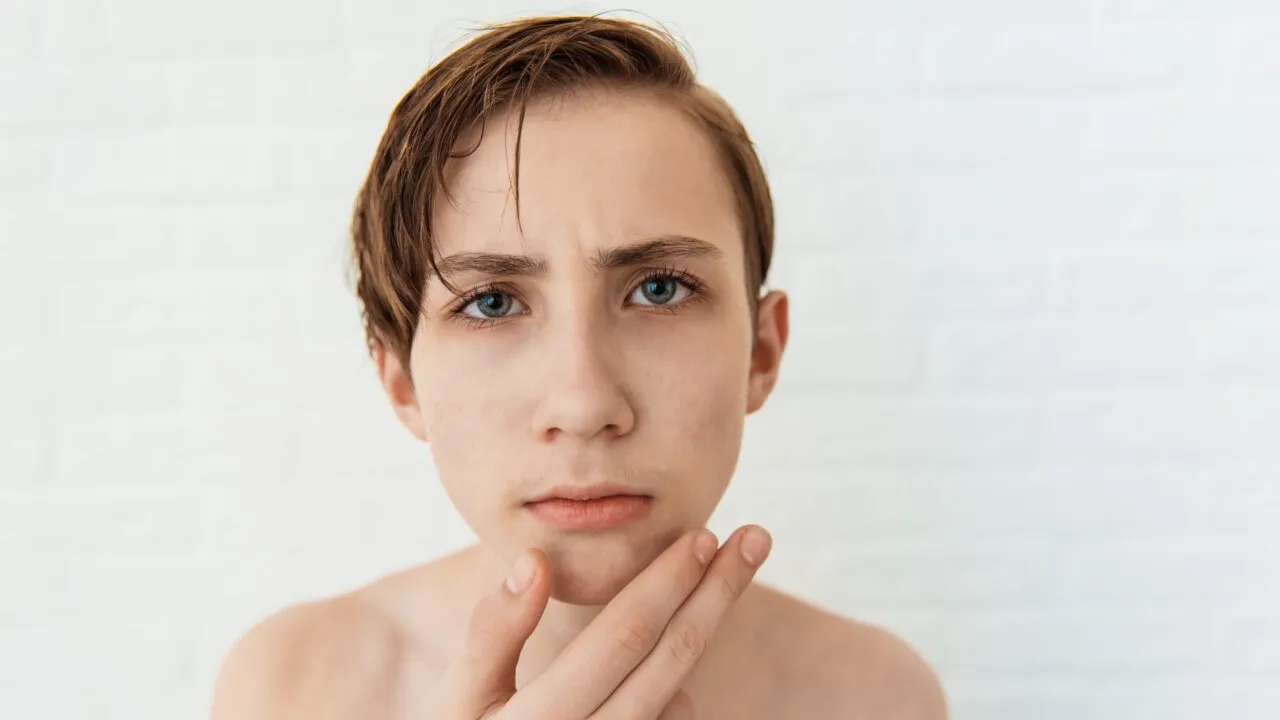 En teenagedreng med blå øjne og vådt, sideskudt brunt hår kigger tæt på kameraet, holder hagen med den ene hånd og ser bekymret ud mod en hvid murstensbaggrund.