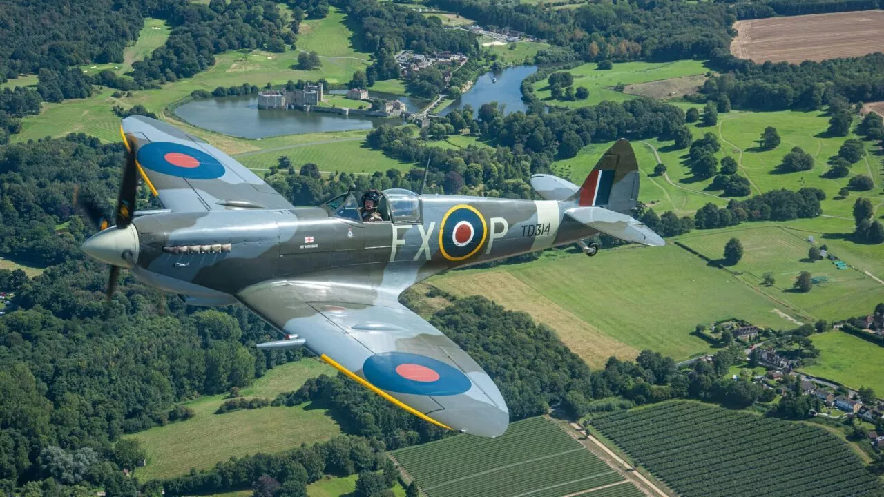 Et gammelt britisk Spitfire-jagerfly fra Anden Verdenskrig med RAF-rundeller flyver over et frodigt grønt landskab med marker, træer og en fjern sø med en bygning på bredden.