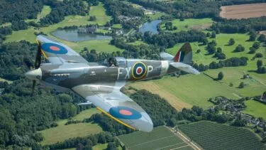 Et gammelt britisk Spitfire-jagerfly fra Anden Verdenskrig med RAF-rundeller flyver over et frodigt grønt landskab med marker, træer og en fjern sø med en bygning på bredden.