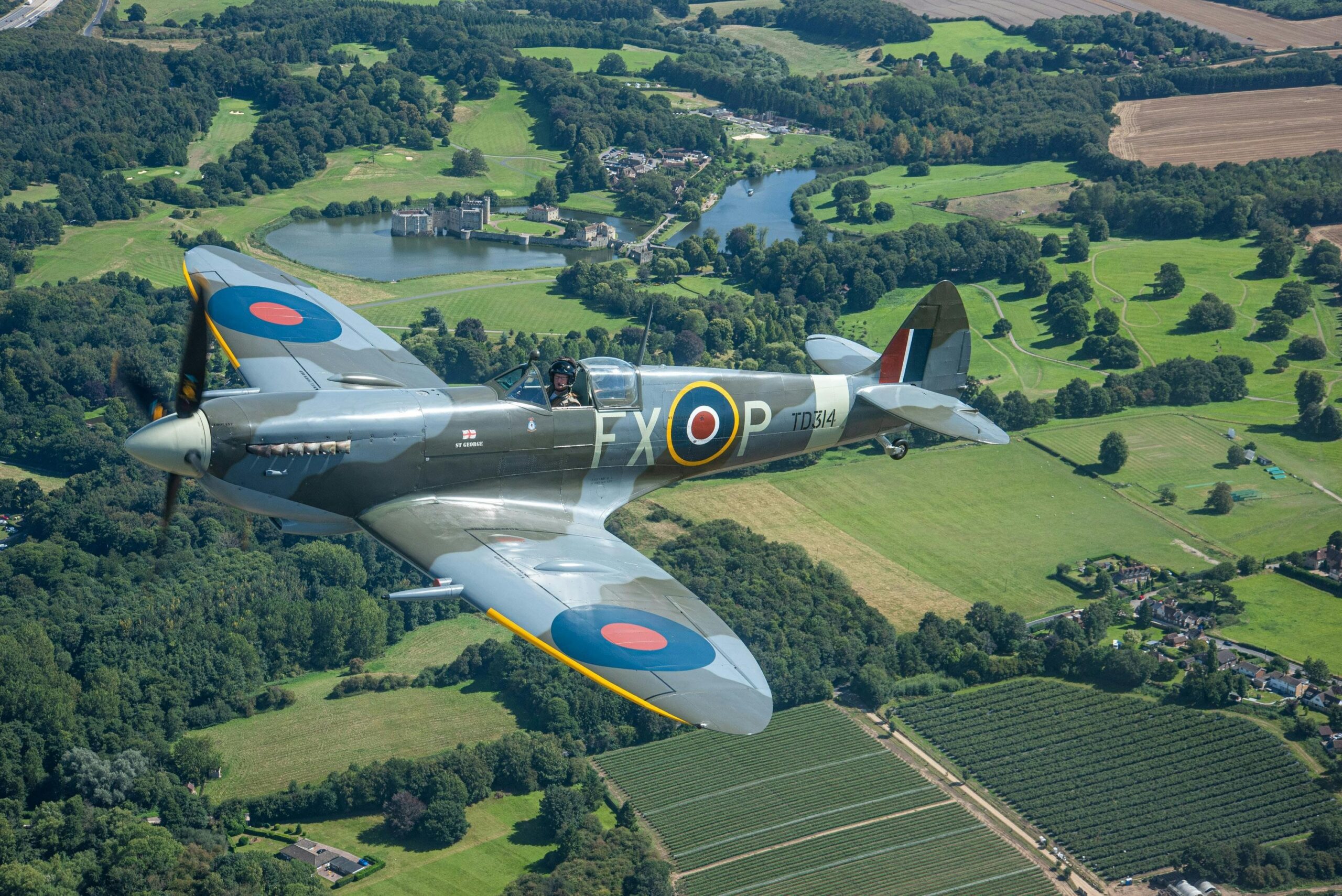 Et gammelt britisk Spitfire-jagerfly fra Anden Verdenskrig med RAF-rundeller flyver over et frodigt grønt landskab med marker, træer og en fjern sø med en bygning på bredden.