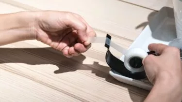 En person trækker en stribe klar tape ud af en sort tapedispenser på et lyst træbord.