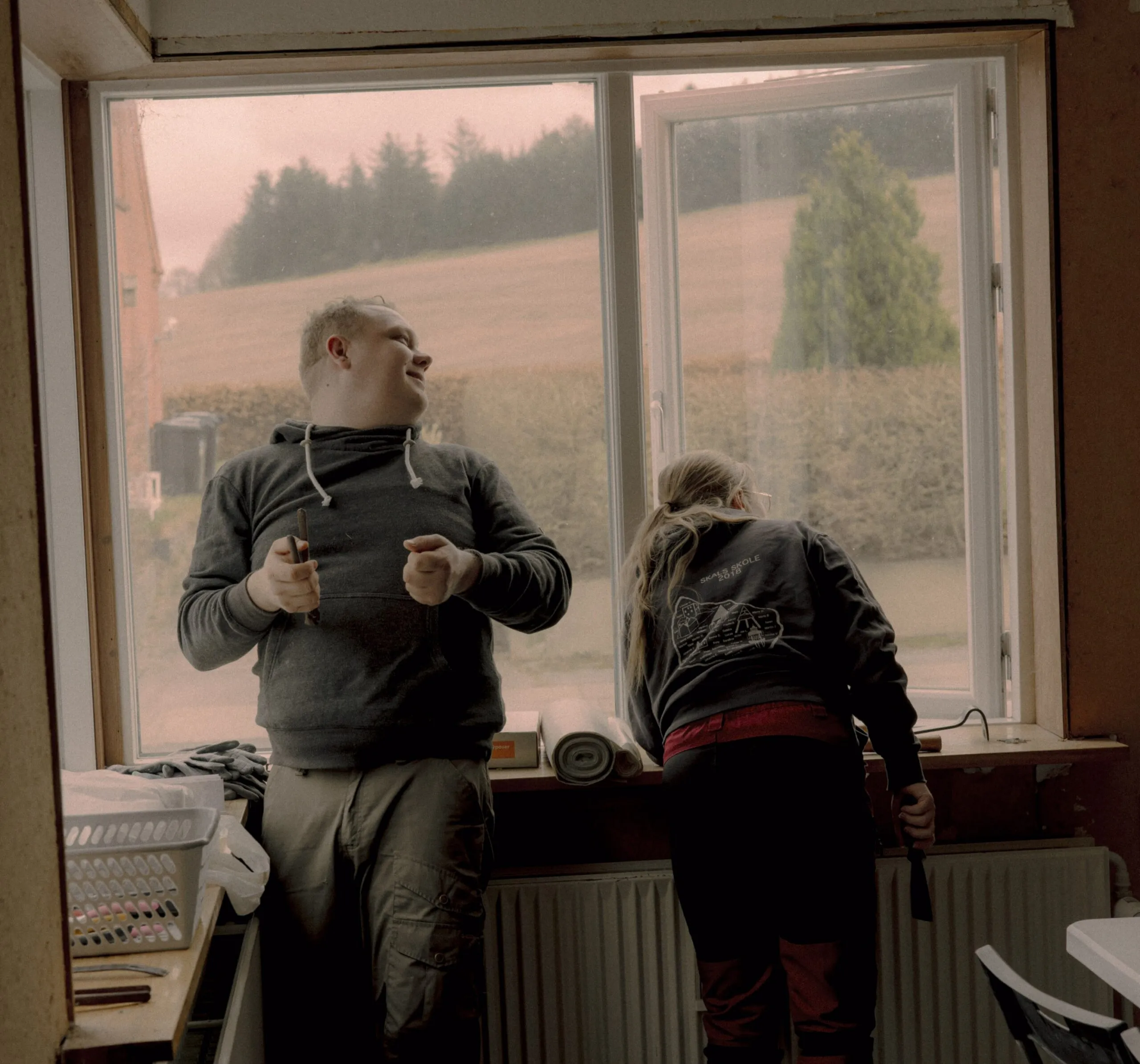 To mennesker står ved et åbent vindue; den ene smiler og holder et værktøj, mens den anden læner sig frem og arbejder. Udenfor kan man se marker og træer under en overskyet himmel. I vindueskarmen ligger forskelligt værktøj og materialer.