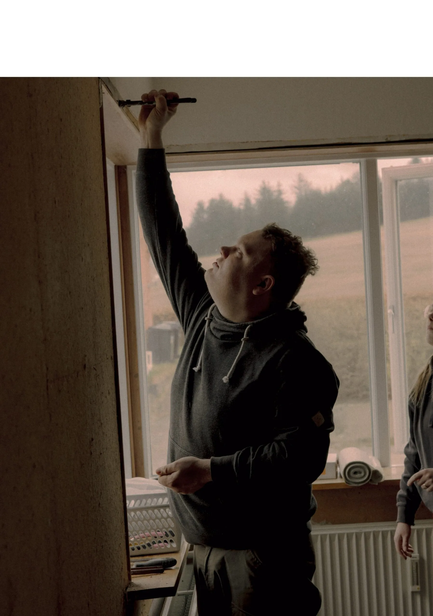 En person står på tæer og maler toppen af en væg nær et vindue, mens en anden person står i nærheden og ser på. Rummet ser ud til at være under renovering, og man kan se værktøj og et bord.