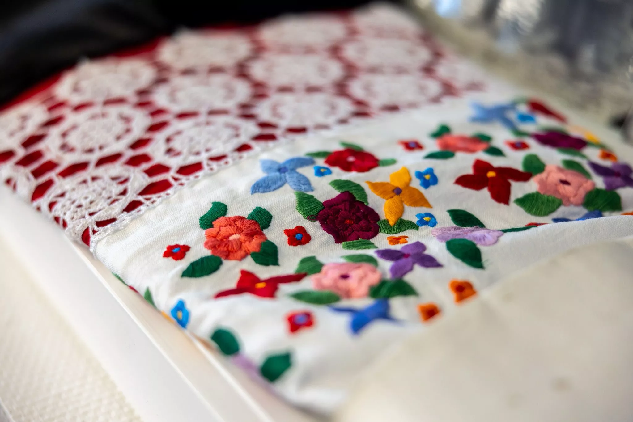 Nærbillede af et stykke hvidt stof broderet med farverige blomster og blade, ved siden af et stykke hvid blonde med et cirkulært mønster på en rød baggrund.
