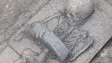 Et delvist udgravet menneskeskelet i en arkæologisk udgravning med en stor metalklinge placeret på tværs af halsen og armene, indlejret i jorden.
