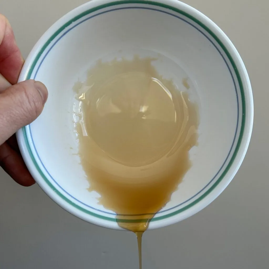 En hånd holder en hvid skål med grønne striber, der er vippet, så honning eller sirup samler sig i bunden og drypper ud af skålen.
