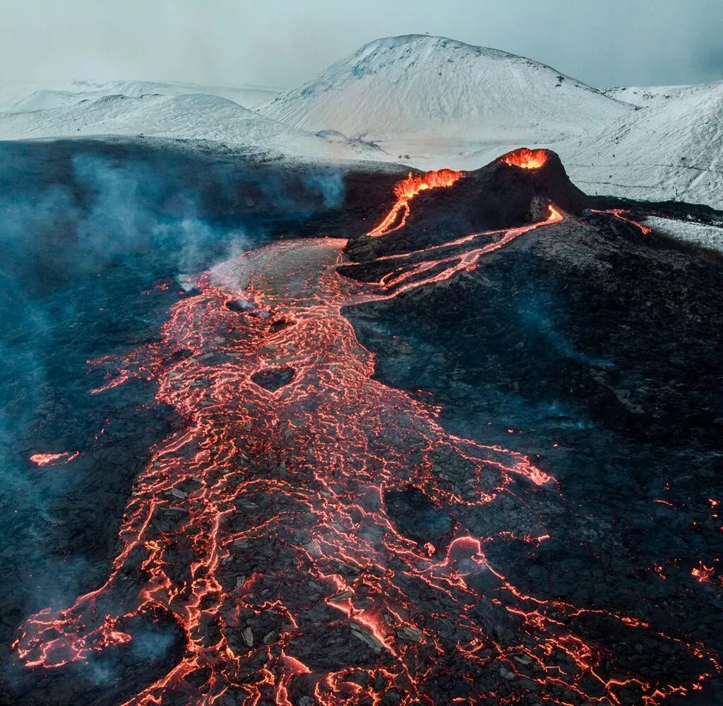 En flod af lysende, glødende lava flyder fra en vulkan i udbrud og skærer sig gennem mørke vulkanske klipper med snedækkede bjerge i baggrunden og røg, der stiger op i himlen.