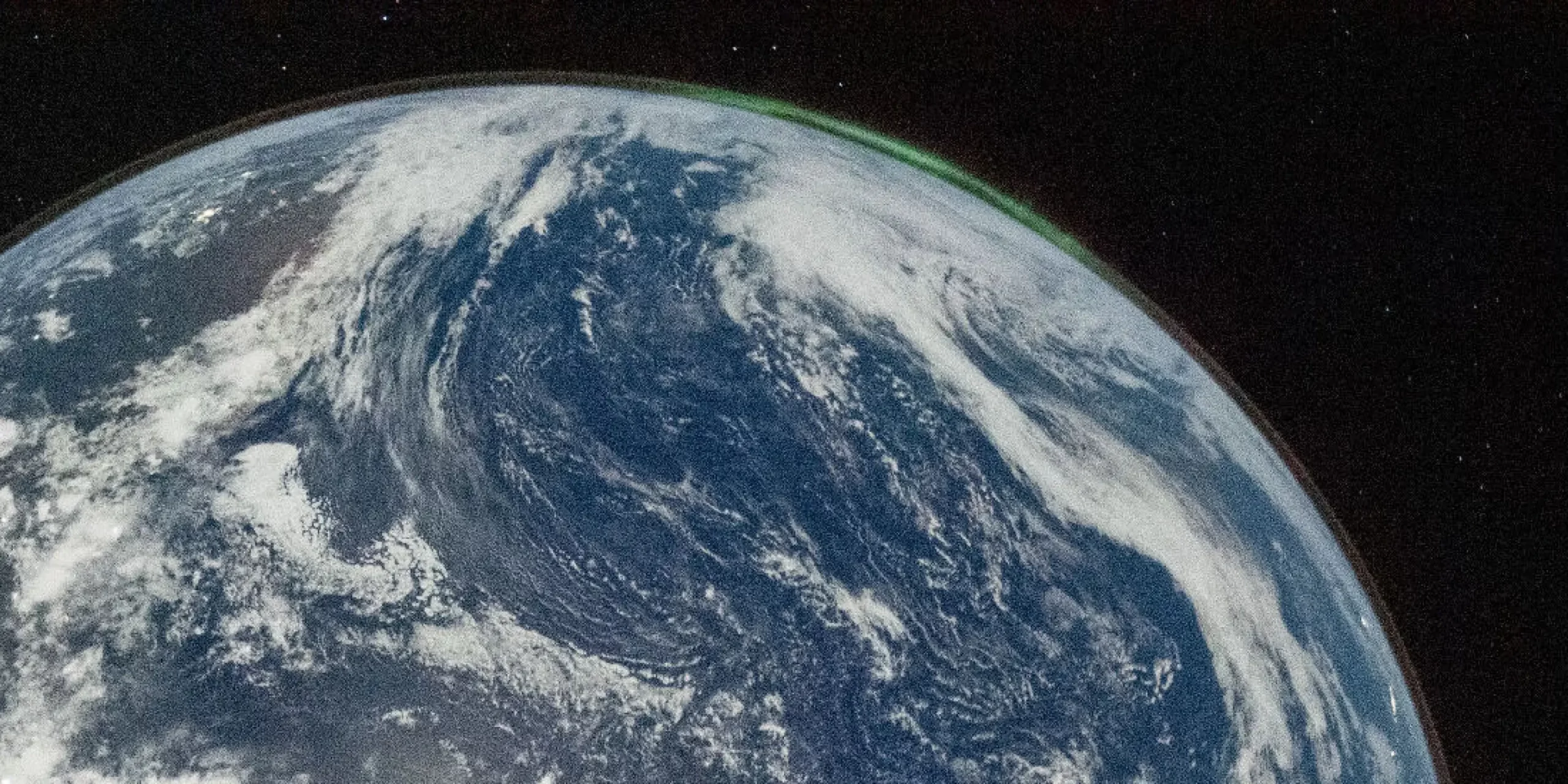 Et billede af Jorden fra rummet viser hvirvlende hvide skyer over blå oceaner. Et svagt grønt nordlys er synligt langs den buede kant af planeten, med det ydre rums mørke i baggrunden.