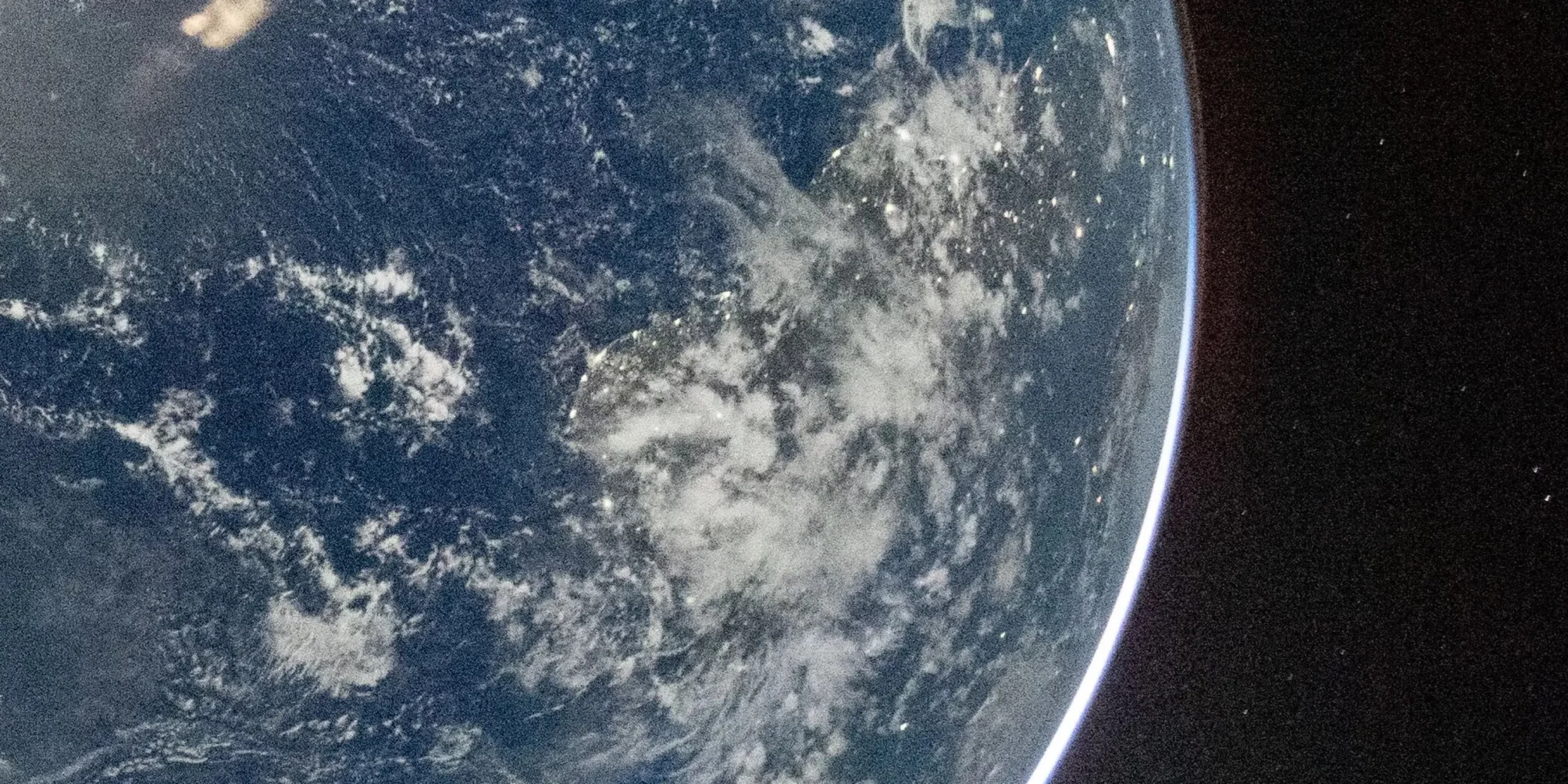 Udsigt over Jorden fra rummet, der viser en buet kant af planeten med blå oceaner, spredte hvide skyer og nogle synlige landmasser under skydækket. Baggrunden er det mørke rum.
