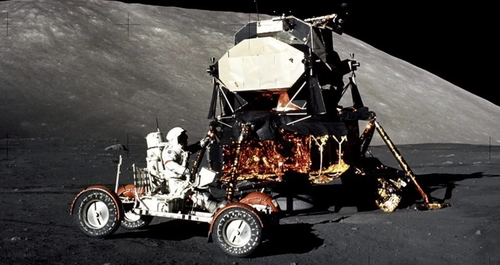En astronaut i rumdragt kører en månerover i nærheden af et guldfoliebeklædt månemodul på månens stenede overflade med en stor bakke eller højderyg i baggrunden under en sort himmel.