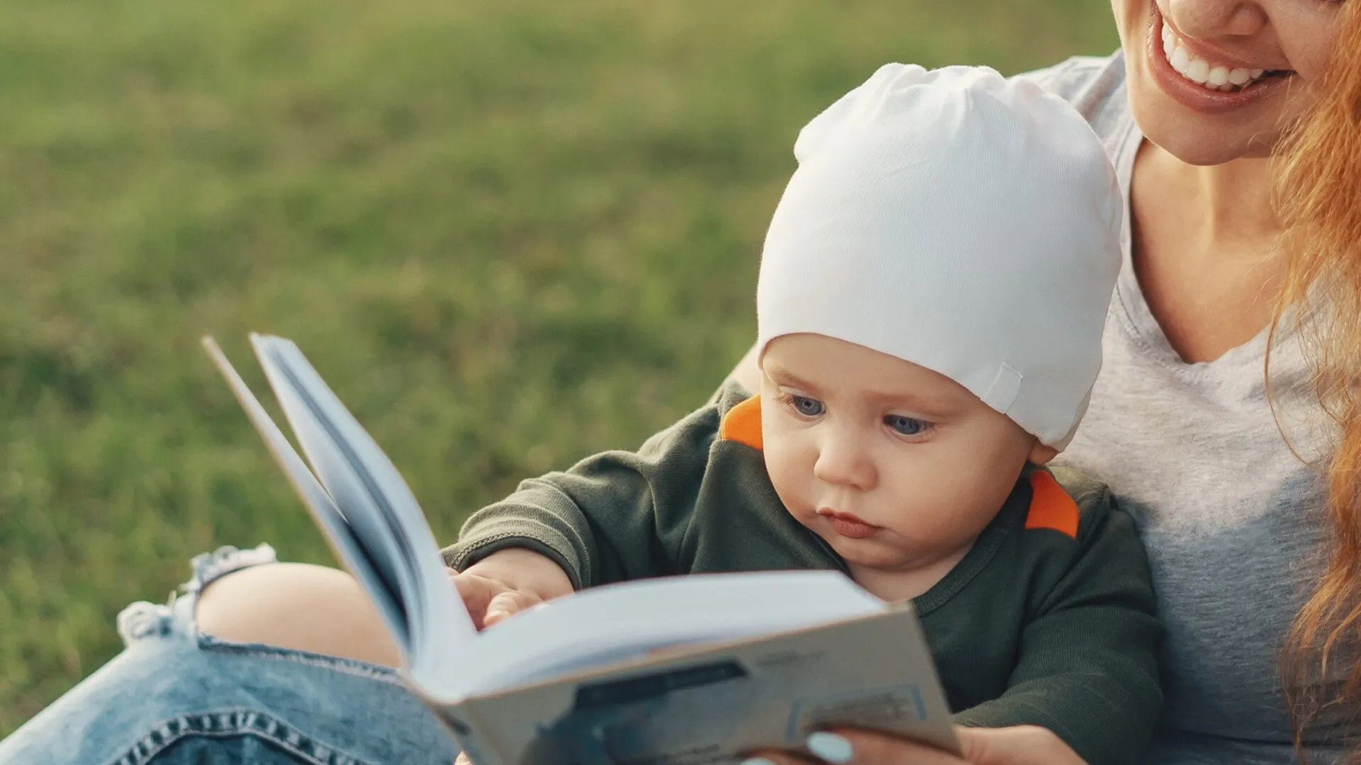 En baby med en hvid hue sidder på skødet af en kvinde udendørs, mens de begge kigger i en åben bog. Kvinden smiler, og de sidder på græsset i naturligt lys.