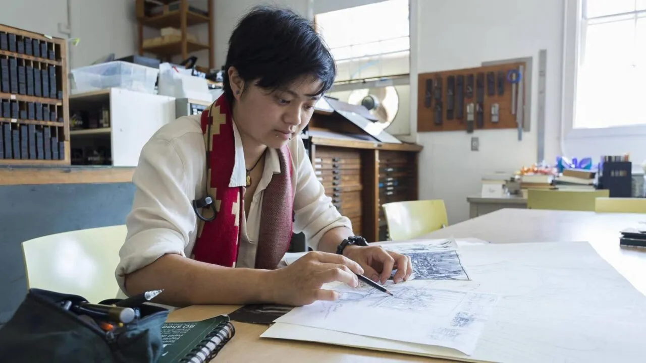 En person med kort mørkt hår sidder ved et skrivebord i et lyst atelier og tegner arkitekttegninger på store ark papir med blyant og lineal. Omkring dem er der kunstartikler og værktøj.