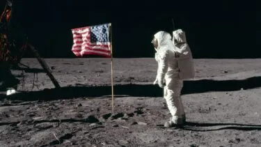 En astronaut i en hvid rumdragt står på Månens overflade ved siden af et amerikansk flag med en månelandingsfartøj delvist synligt i baggrunden. Jorden er grå og dækket af fodspor under en sort himmel.