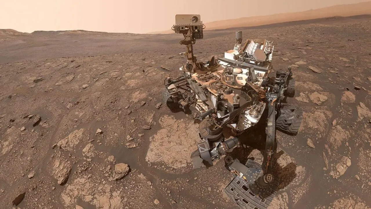 En Mars-robot med kameraer og videnskabelige instrumenter står på en stenet, rødlig Marsoverflade under en diset himmel, omgivet af fjerne bakker.