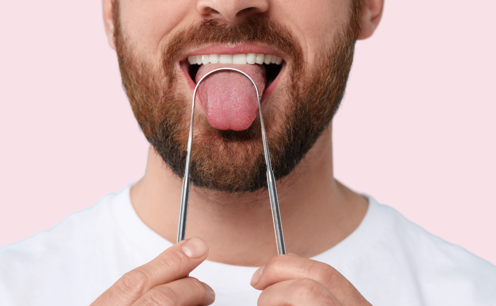 En mand med skæg bruger en tungeskraber af metal på sin udspilede tunge, mens han holder værktøjet med begge hænder mod en lyserød baggrund.