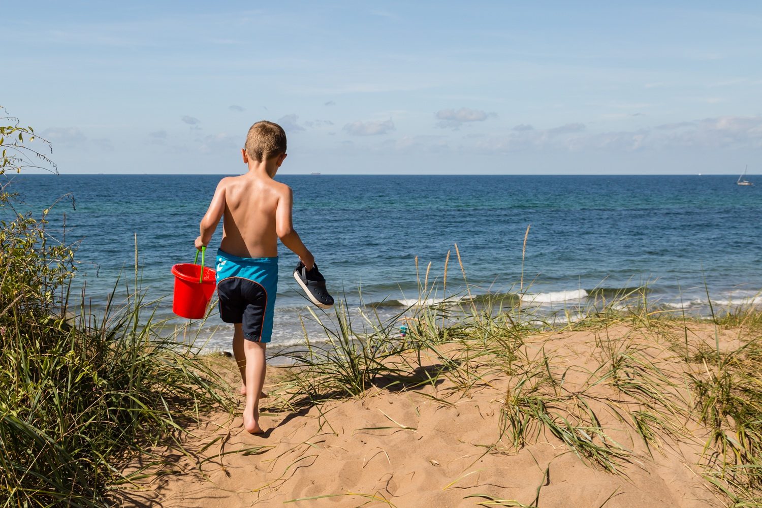 En ung dreng iført blå badebukser går ned ad en sandklit mod havet med en rød spand i den ene hånd og en sko i den anden, med blå himmel og bølger i baggrunden.